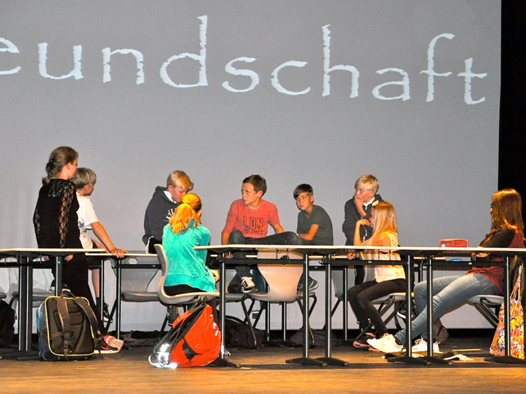 Eine Szene an Schultischen vor einer Projektion mit dem Wort „Freundschaft“.